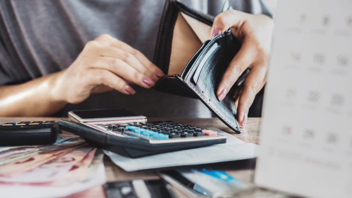 Una mujer revisa su cartera vacía mientras se encuentra sentada en una mesa llena de billetes, recibos y una calculadora. Su expresión y postura reflejan preocupación por su situación financiera. Frente a ella, un teléfono móvil y un calendario sugieren que está intentando negociar deudas o planificar sus pagos.