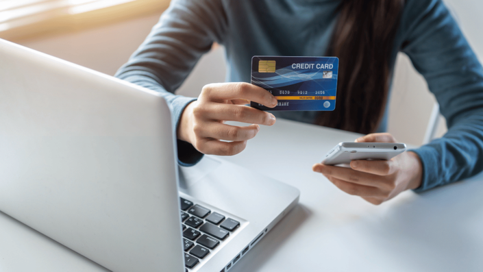 Mujer sosteniendo una tarjeta de crédito sin anualidad mientras usa su teléfono móvil y una computadora portátil. Representa pagos en línea y compras seguras sin costos adicionales.