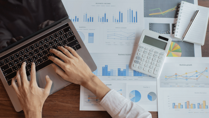 Una persona escribiendo en una laptop sobre documentos financieros con gráficos y estadísticas. Se observan una calculadora, un cuaderno y un bolígrafo, lo que sugiere análisis y planificación económica. Los títulos valores pueden ser clave en la gestión financiera y el crecimiento empresarial.