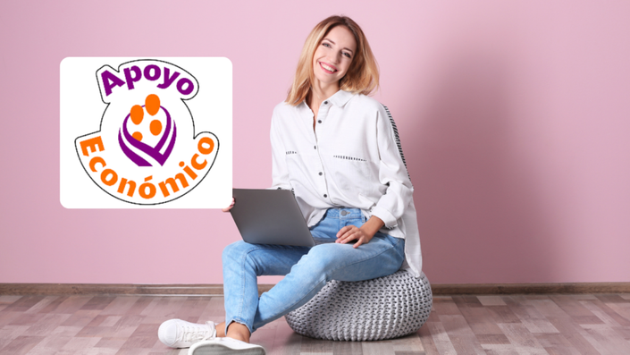 Mujer joven con cabello rubio, sonriendo y sosteniendo una laptop mientras se sienta sobre un puf gris en una habitación con una pared de color rosa. Lleva una camisa blanca y jeans azules